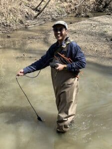 A man wearing a hat, sunglasses, and waders uses a handheld water quality sonde to measure water quality in a murky stream