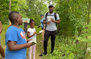 A young man stands on a trail in a bright green forest, talking to kids carrying notebooks.