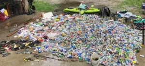 A volunteer in a green kayak sits in front of a giant heap of trash and cans along the banks of a river